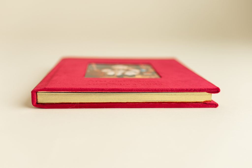 Holiday photo album laying closed in side angle view, highlighting the glinting page edges and red velvet wraparound fabric.