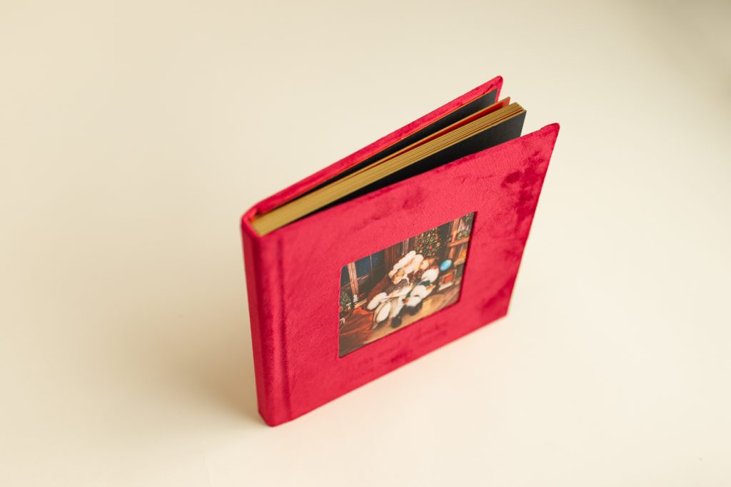 Red velvet photo album standing upright, top angle view showing glint-edged pages; square cover photo features holiday photo.