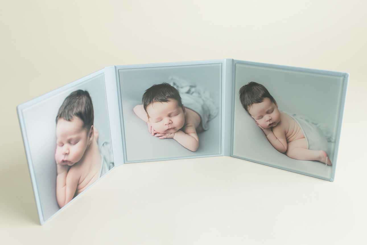 Triplex photo folio open and spread out; features newborn boy portraits with acrylic finish and soft blue fabric cover.