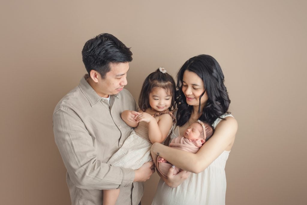 Studio family portrait of parents holding newborn and toddler sibling; all wearing neutral clothing and looking down at baby.